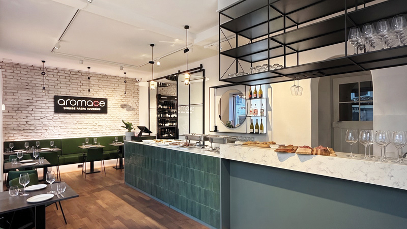 Modernes Restaurantinterieur mit Bar, offenen Regalen, grünen Sitzbereichen und stilvoller Beleuchtung.